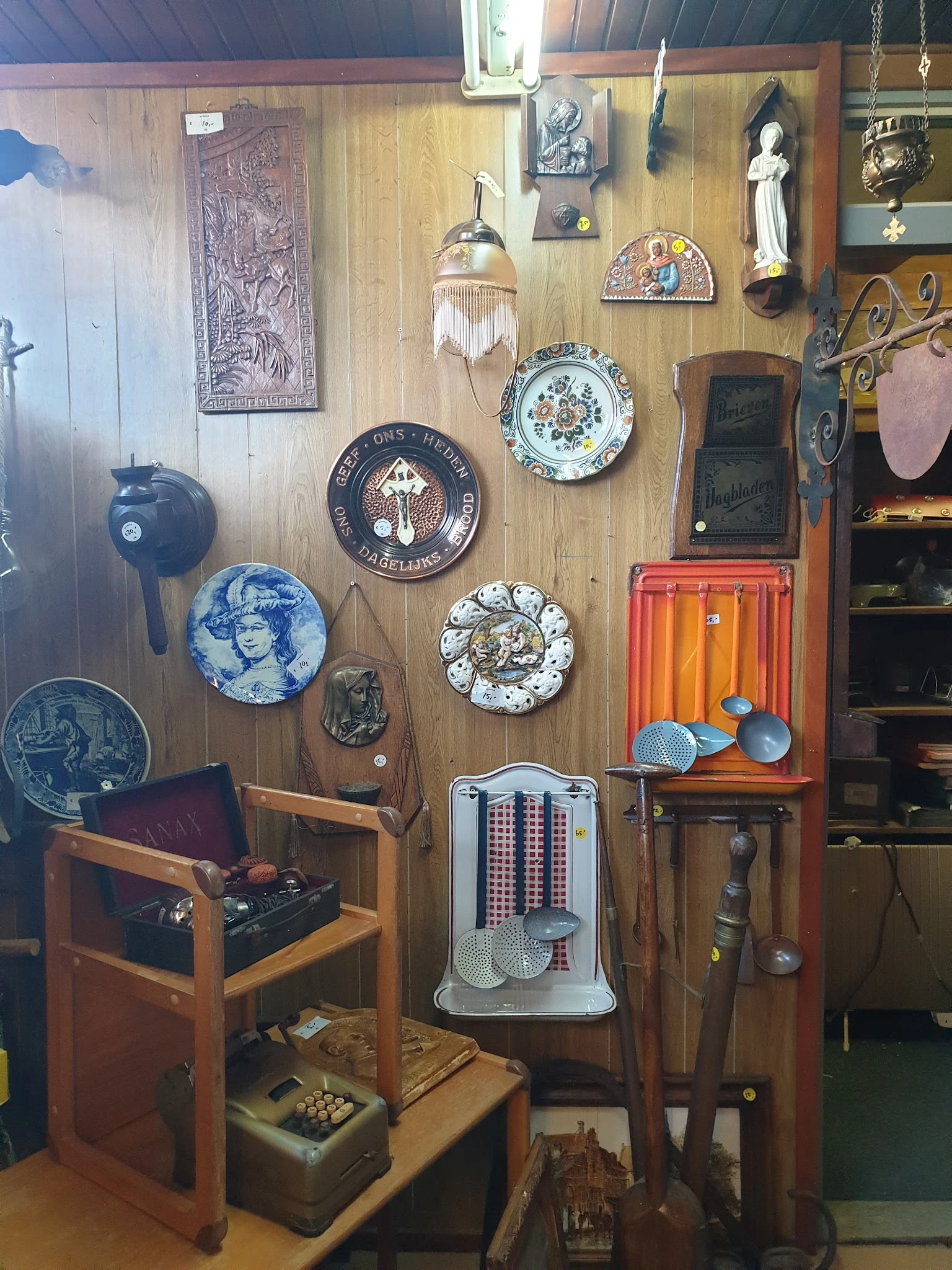 Verzameling tweedehands huisraad aan muur: decoratieve borden, vintage radio, stoelen, spiegel en diverse antieke voorwerpen