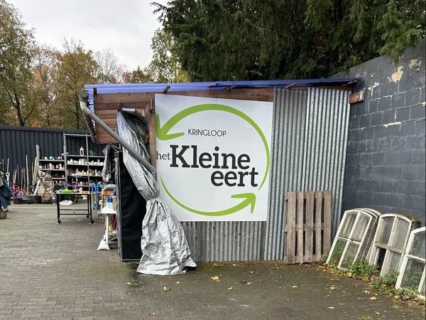 Voorkant van kringloopwinkel met blauwe afdak en logo van Kringloop het Kleine Eert op golfplaten wand