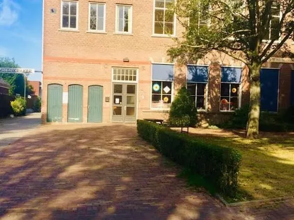 Voorgevel van bakstenen kringloopwinkel met groene deuren en ramen, patio met boom en struiken