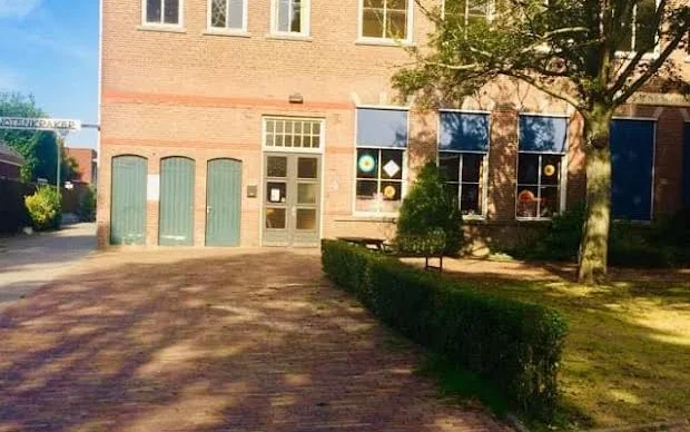 Voorgevel van bakstenen kringloopwinkel met groene deuren en ramen, patio met boom en struiken