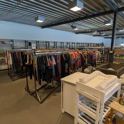 Kringloopwinkel interieur met kledingrekken, witte kasten met huishoudelijke items en open ruimte onder golfplaten plafond