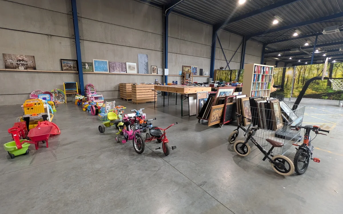 Grote hal met kinderfietsjes, kinderwagens, boekenrekken en diverse spullen uitgestald op betonvloer