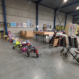 Grote hal met kinderfietsjes, kinderwagens, boekenrekken en diverse spullen uitgestald op betonvloer