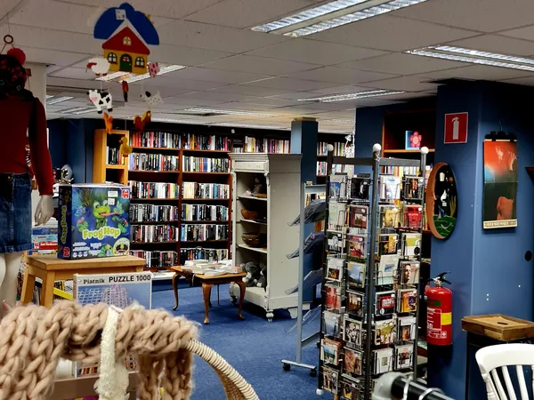 Binnenkant kringloopwinkel met boekenkasten, speelgoed, puzzels en diverse tweedehandsartikelen onder fluorescente verlichtin