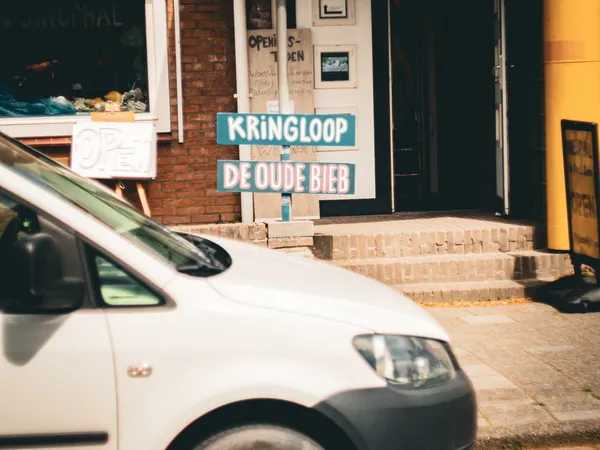 Voorgevel van kringloopwinkel met blauwe uithangbord 'KRINGLOOP DE OUDE BIEB' en witte bestelwagen geparkeerd voor de ingang