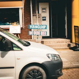 Voorgevel van kringloopwinkel met blauwe uithangbord 'KRINGLOOP DE OUDE BIEB' en witte bestelwagen geparkeerd voor de ingang