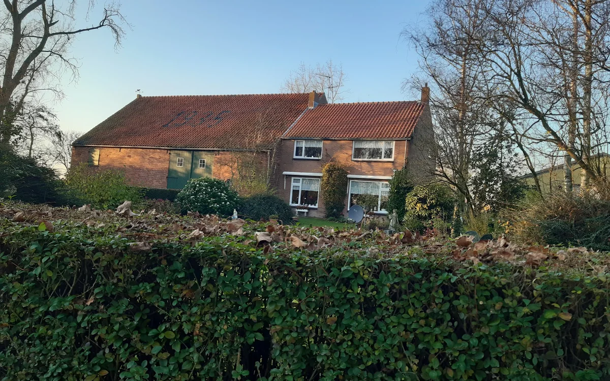 Voorkant van een bakstenen woonhuis met rode pannendaken, omgeven door groen struikgewas en kale bomen
