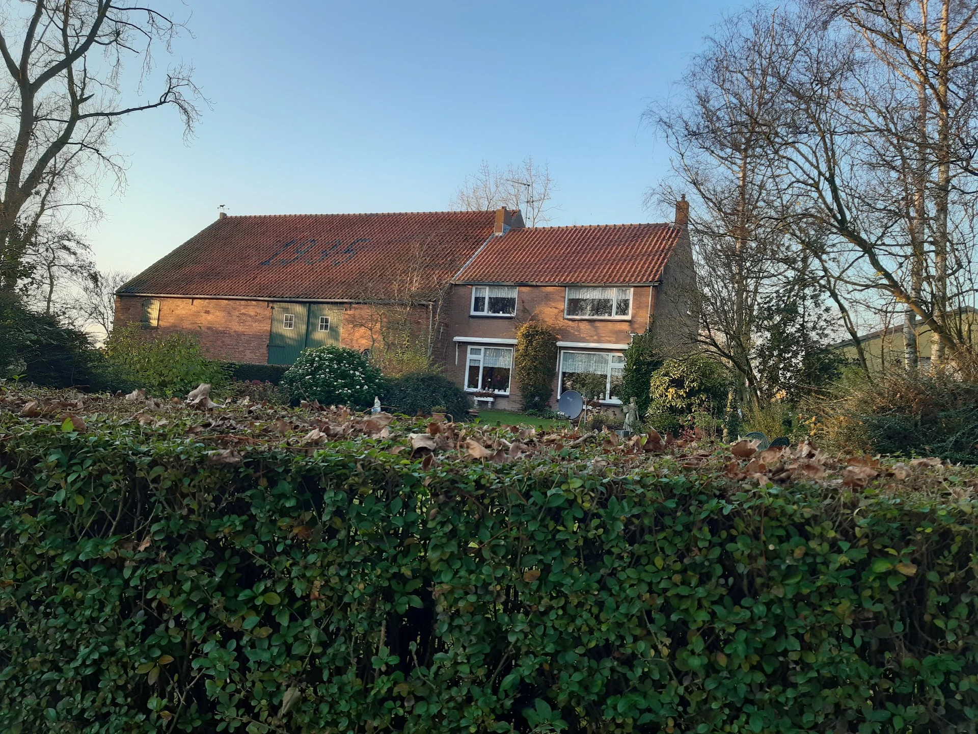 Voorkant van een bakstenen woonhuis met rode pannendaken, omgeven door groen struikgewas en kale bomen