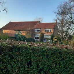 Voorkant van een bakstenen woonhuis met rode pannendaken, omgeven door groen struikgewas en kale bomen