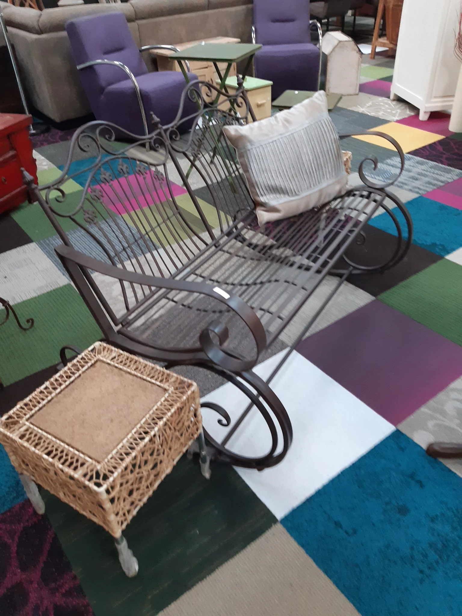 Tweedehands meubels en huishoudartikelen op een gekleurde vloer, waaronder een metalen stoel met krullen en een rieten mand