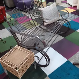 Tweedehands meubels en huishoudartikelen op een gekleurde vloer, waaronder een metalen stoel met krullen en een rieten mand