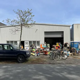 Voorgevel van kringloopwinkel met grote grijze deuren, daarvoor uitgestalde tweedehands spullen en een auto geparkeerd