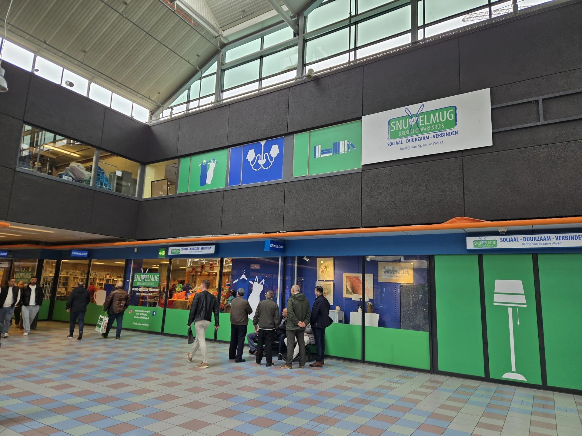 Winkelinterieur met blauwe en groene gevels, stellingen met spullen en bezoekers in de kringloopwinkel