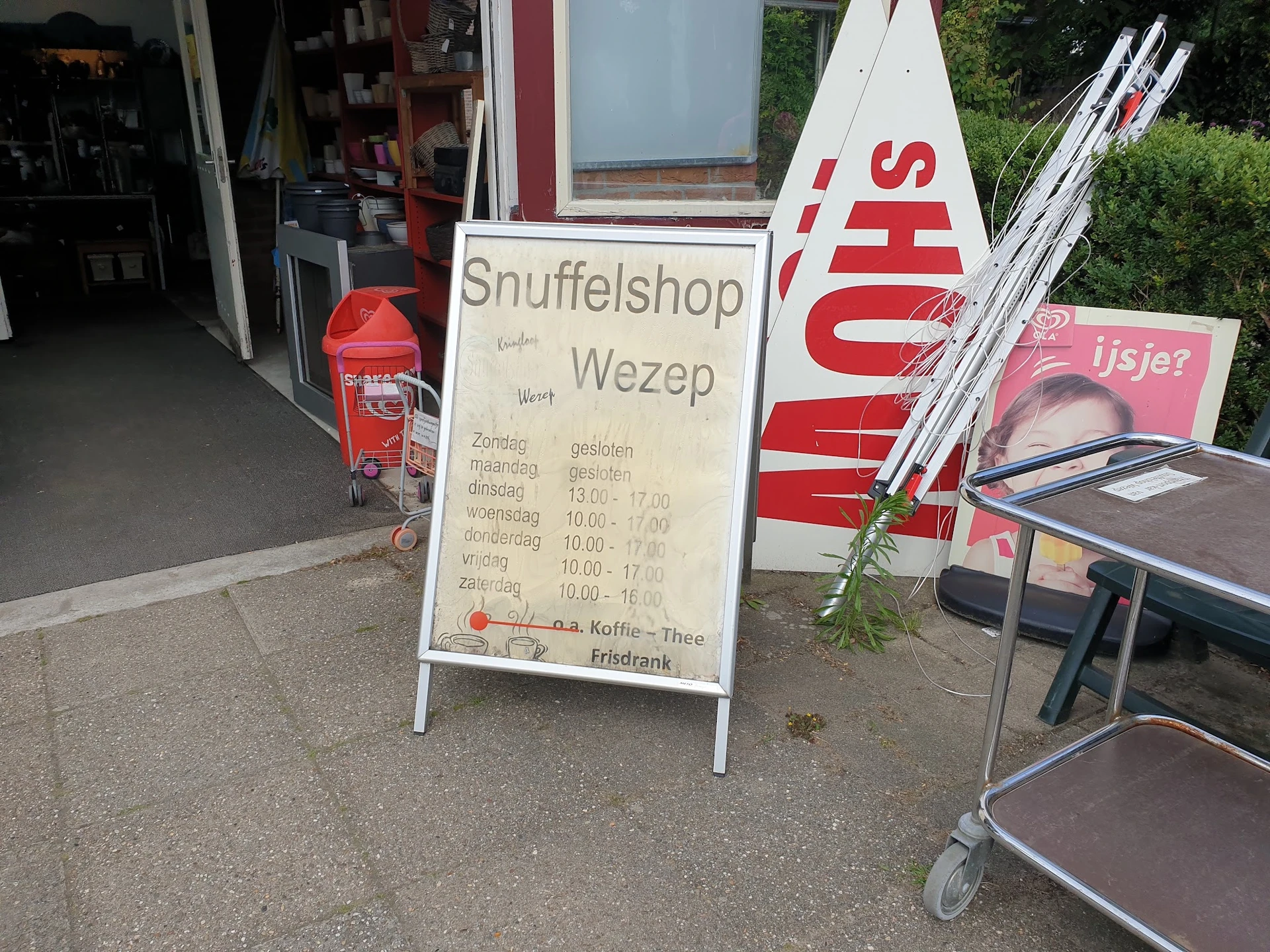 Voorkant van kringloopwinkel met uithangbord en reclameborden, deels belemmerd door voorwerpen