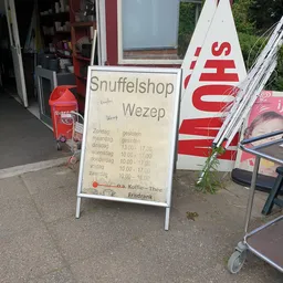 Voorkant van kringloopwinkel met uithangbord en reclameborden, deels belemmerd door voorwerpen