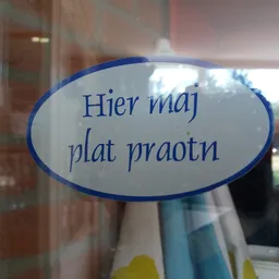 Oval blauw bord met Nederlandse tekst 'Hier maj plat praotn' op een winkelruit