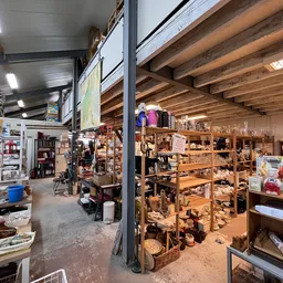Ruim interieur van kringloopwinkel met houten stellingen, diverse spullen, en expositieruimte onder houten balkenplafond