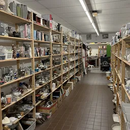 Lange gang tussen houten stellingen vol met huishoudelijke spullen, serviesgoed en diversen in een kringloopwinkel