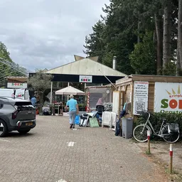 Voorkant van kringloopwinkel met parkeerplaats, fietsen en terras met parasol