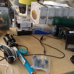 Diverse tweedehands elektronica en accessoires op tafel, waaronder koptelefoon, gereedschap en verpakte items met prijssticke