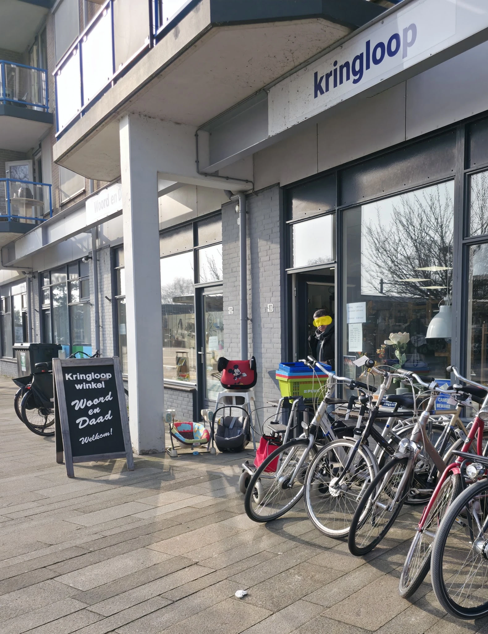 Voorgevel van kringloopwinkel met grote ramen, zwarte deur en fietsen geparkeerd ervoor