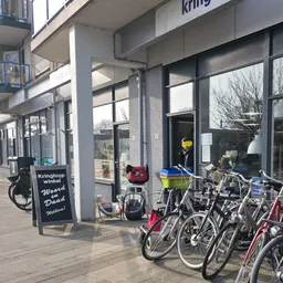 Voorgevel van kringloopwinkel met grote ramen, zwarte deur en fietsen geparkeerd ervoor