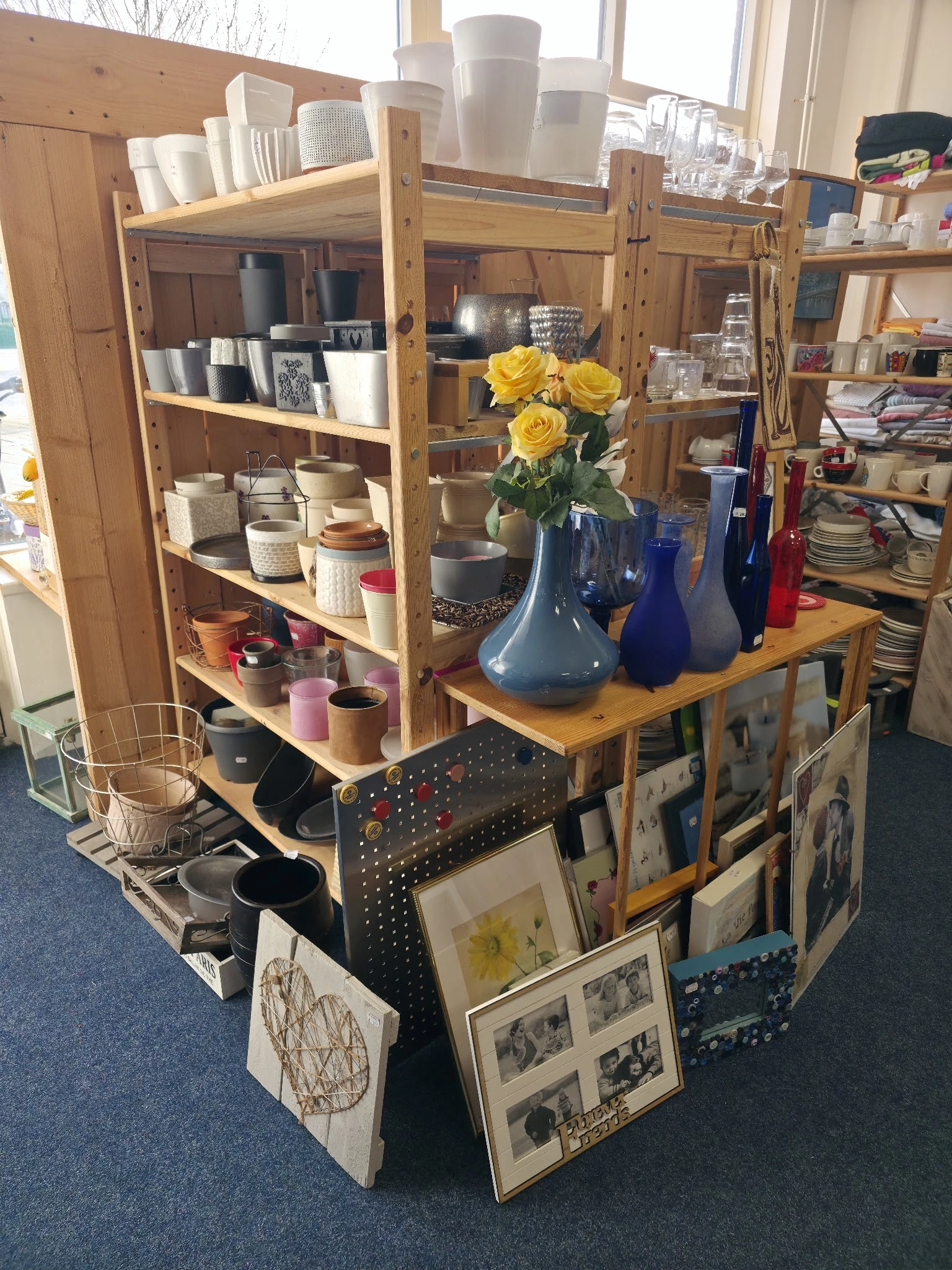 Houten tafel met blauwe vazen, gele bloemen en diverse huishoudartikelen, kasten met servies en decoratieve spullen op achter