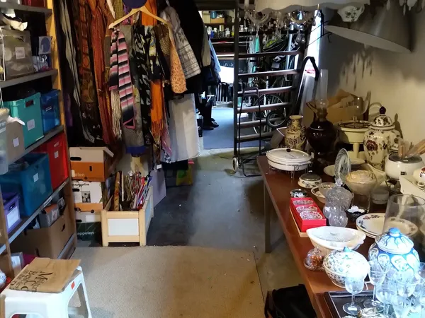 Binnenaanzicht van kringloopwinkel met kledingrekken links, kasten met spullen rechts en tafel met servies voorgrond