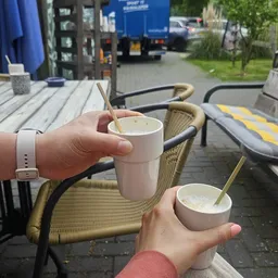 Twee handen houden witte bekers met drank en rietjes op een terras