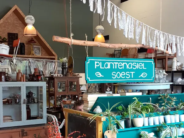 Winkelinterieur met houten stellingen, groene muur, neonbord 'Plantengasiel Soest' en plantenuitstalling op blauwe tafel