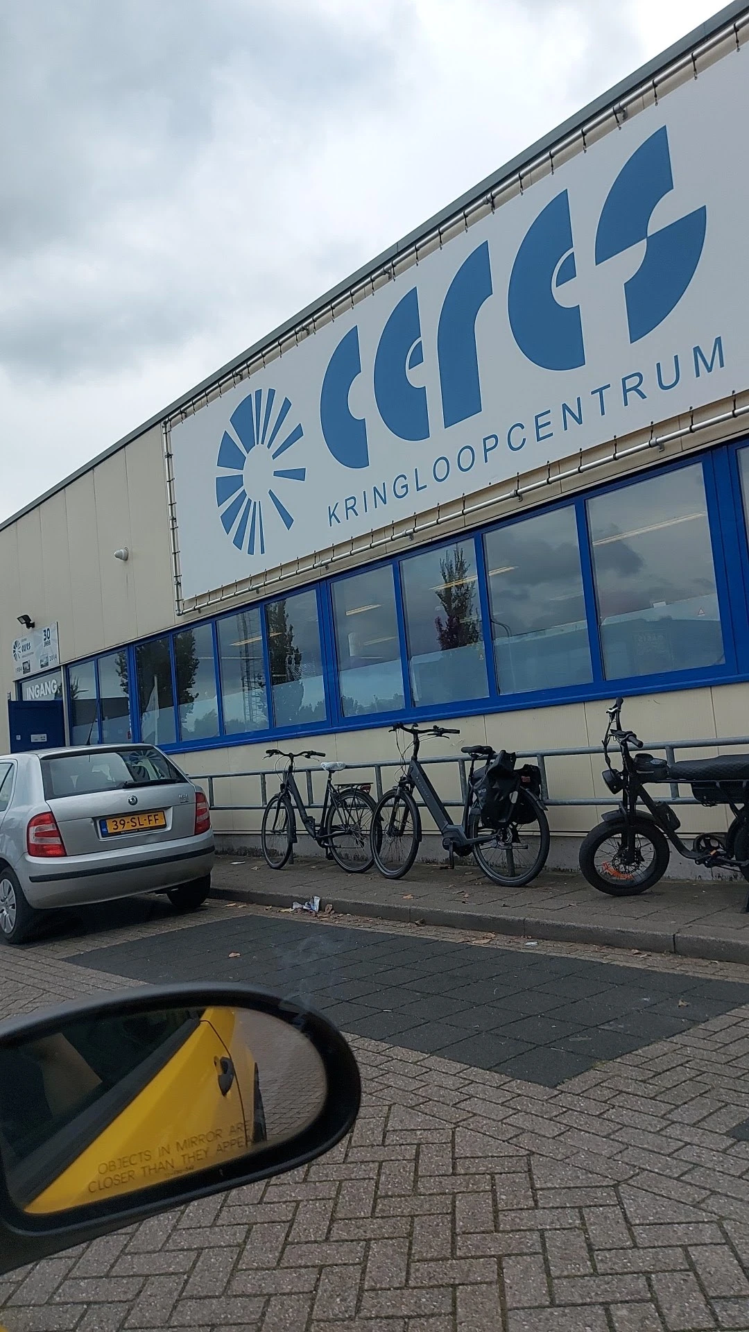 Gevel van kringloopwinkel met blauwe letters en ramen, parkeerplaats met auto's en fietsen ervoor