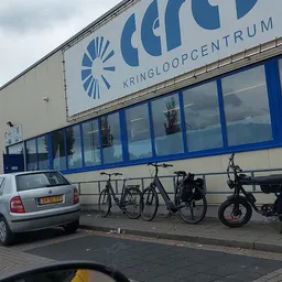 Gevel van kringloopwinkel met blauwe letters en ramen, parkeerplaats met auto's en fietsen ervoor