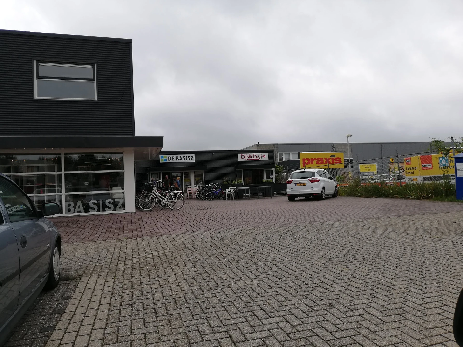 Voorkant van kringloopwinkel met zwart pand, wit deel links met naam 'DE BASISZ' zichtbaar, parkeerplaats met enkele auto's