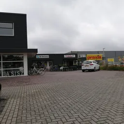 Voorkant van kringloopwinkel met zwart pand, wit deel links met naam 'DE BASISZ' zichtbaar, parkeerplaats met enkele auto's