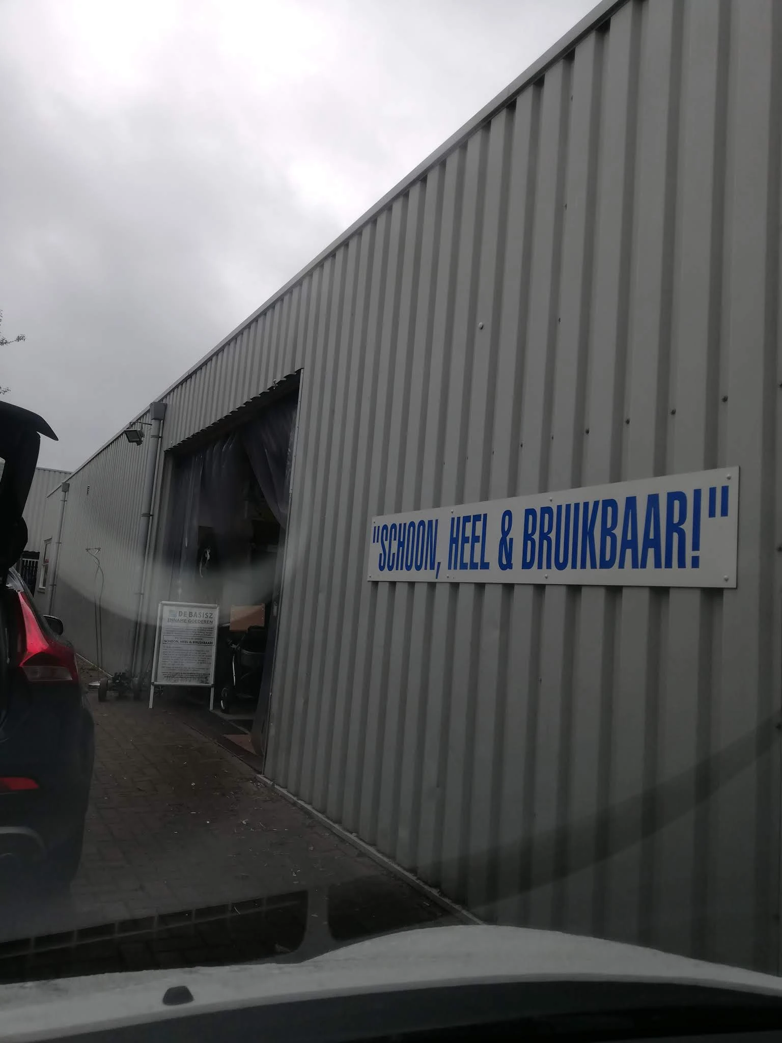 Grijze golfplaten gevel van kringloopwinkel met blauwe tekst 'SCHOON, HEEL & BRUIKBAAR!' en open ingang
