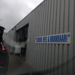 Grijze golfplaten gevel van kringloopwinkel met blauwe tekst 'SCHOON, HEEL & BRUIKBAAR!' en open ingang
