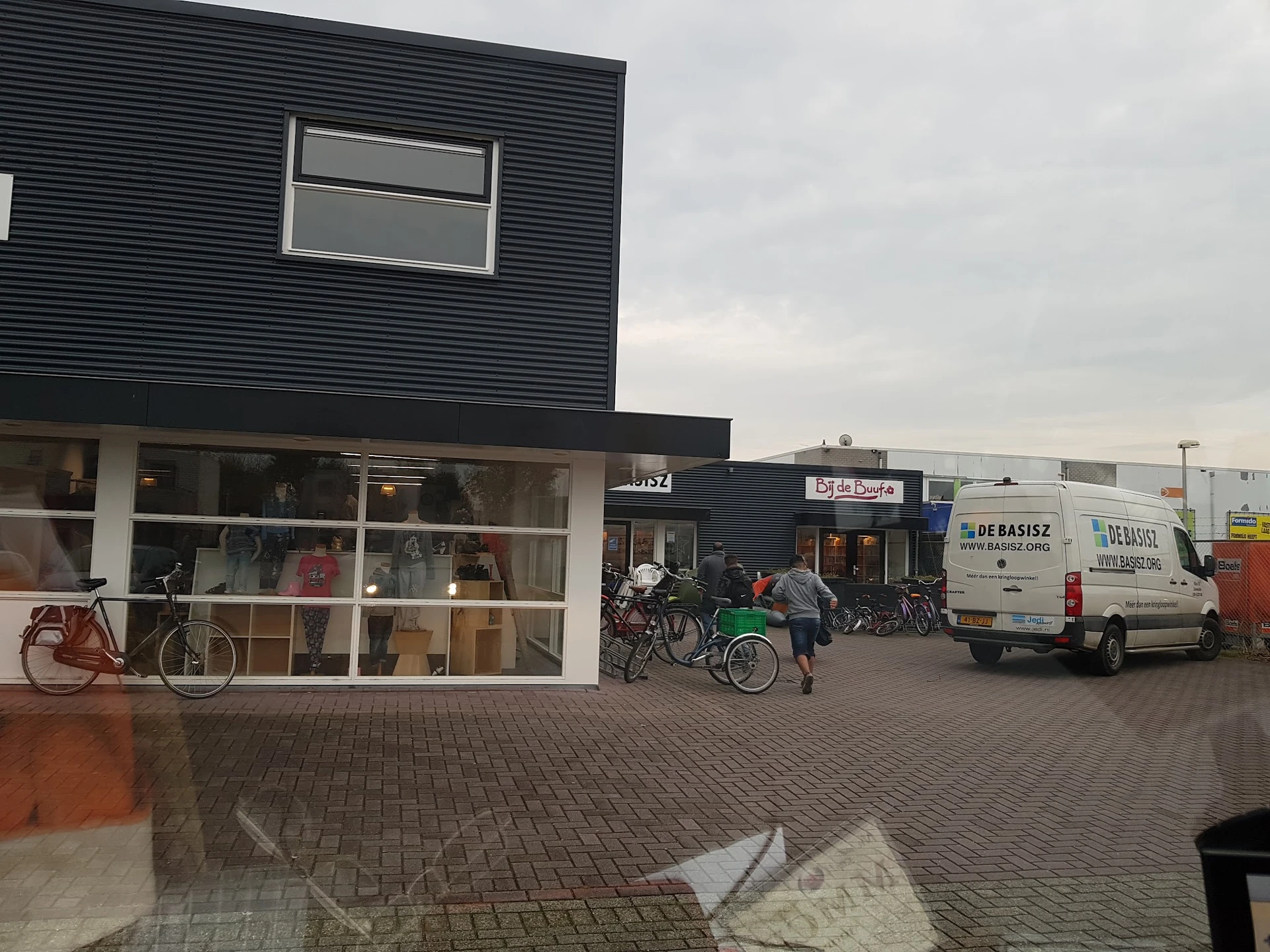 Gevel van kringloopwinkel met donkergrijze golfplaten bovenkant, witte onderkant met grote ramen, fietsen ervoor en parkeerte