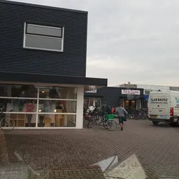 Gevel van kringloopwinkel met donkergrijze golfplaten bovenkant, witte onderkant met grote ramen, fietsen ervoor en parkeerte