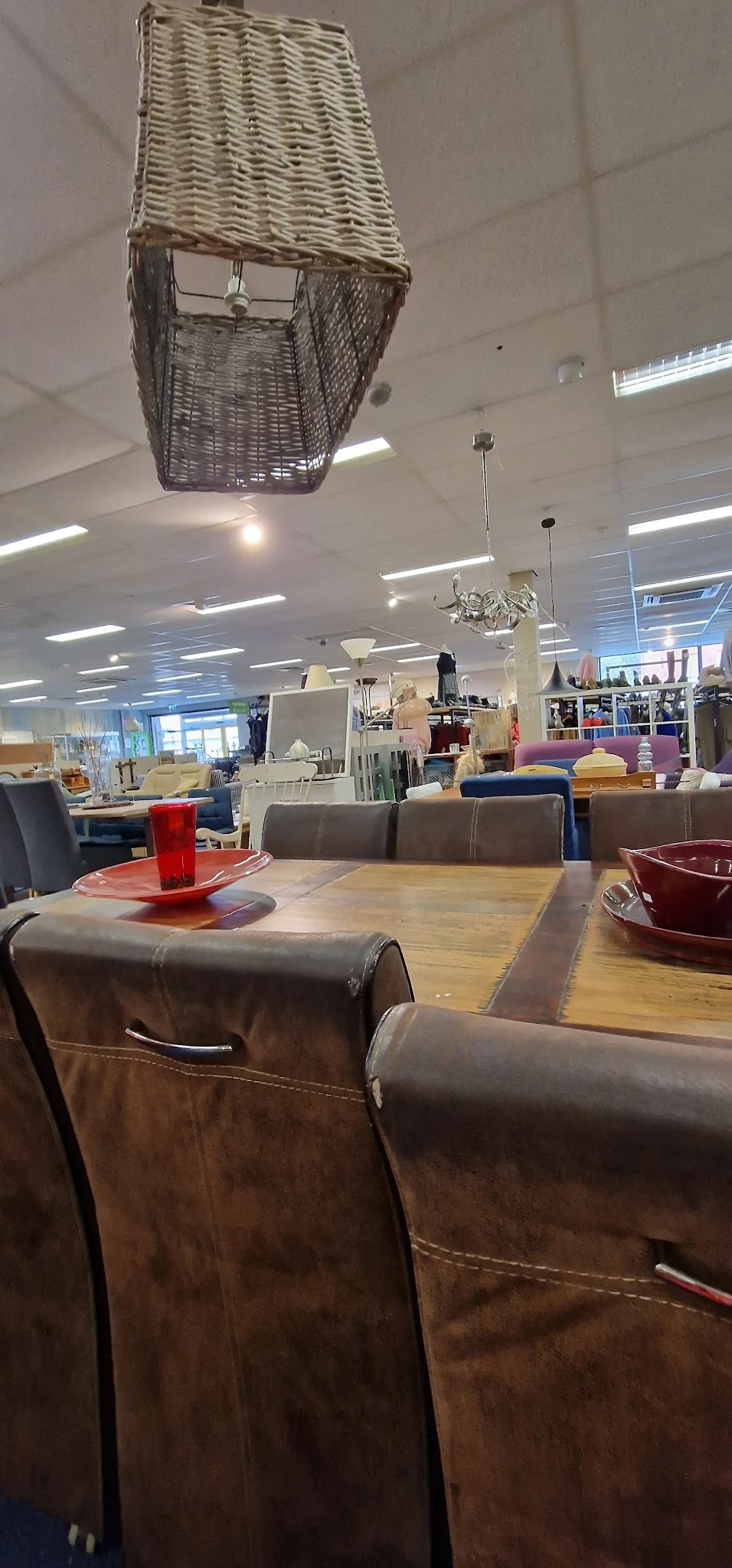 Winkelvloer met houten tafel, bruine stoelen en rode accessoires, stellingen met kleding en huishoudartikelen op de achtergro