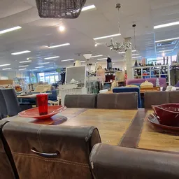 Winkelvloer met houten tafel, bruine stoelen en rode accessoires, stellingen met kleding en huishoudartikelen op de achtergro