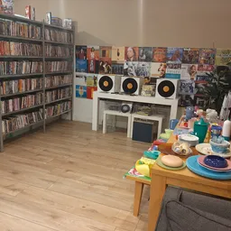 Winkelvloer met cd-kasten, een witte kast met draaitafels, dvd-collectie aan muur, en tafel met serviesgoed en speelgoed