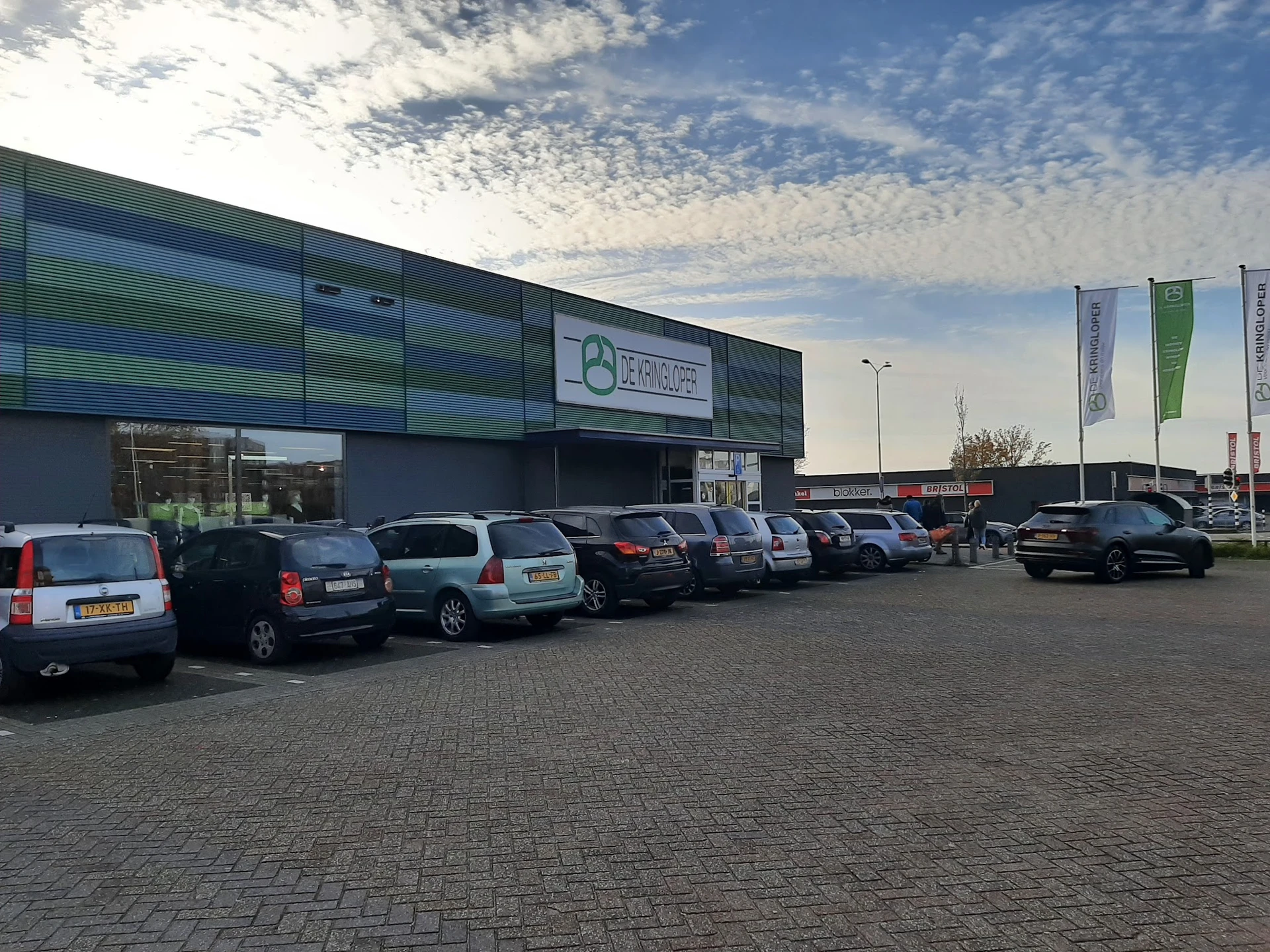 Voorgevel van kringloopwinkel met groene en blauwe panelen, parkeerplaats met auto's ervoor