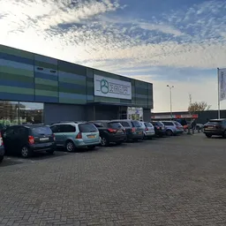 Voorgevel van kringloopwinkel met groene en blauwe panelen, parkeerplaats met auto's ervoor