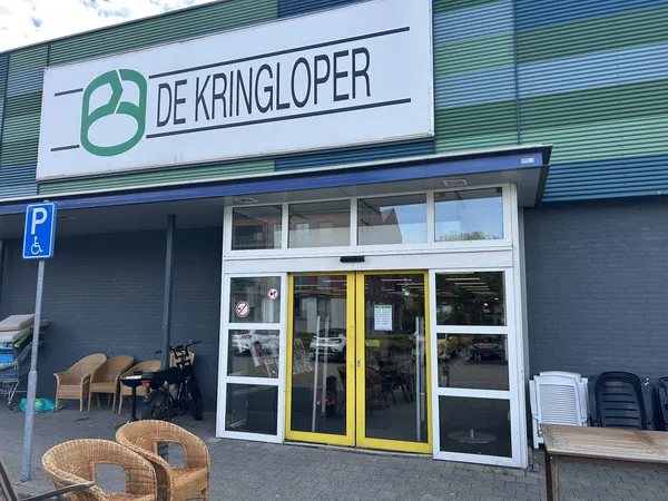 Gevel van kringloopwinkel met groene logo, witte deuzen met gele accenten, en spullen uitgestald op het trottoir