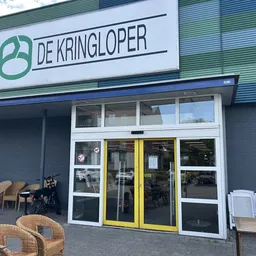 Gevel van kringloopwinkel met groene logo, witte deuzen met gele accenten, en spullen uitgestald op het trottoir