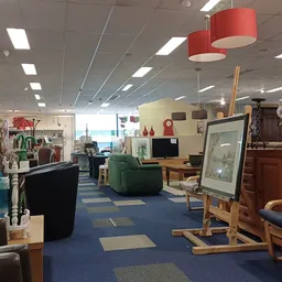 Winkelvloer van kringloopwinkel met meubels, fauteuils, lampen en huisraad uitgestald, blauwe vloer en rode hanglampen.