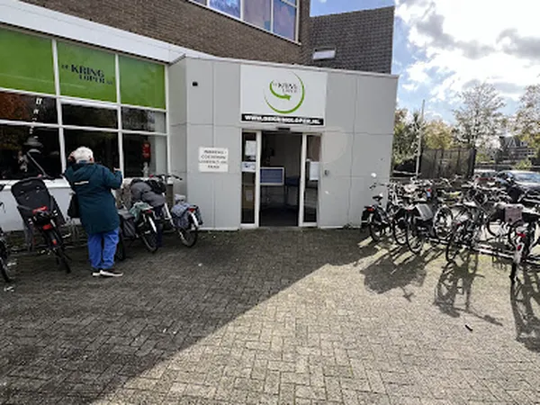 Witte gevel van kringloopwinkel met groene zijvleugel, fietsen en brommers buiten geparkeerd