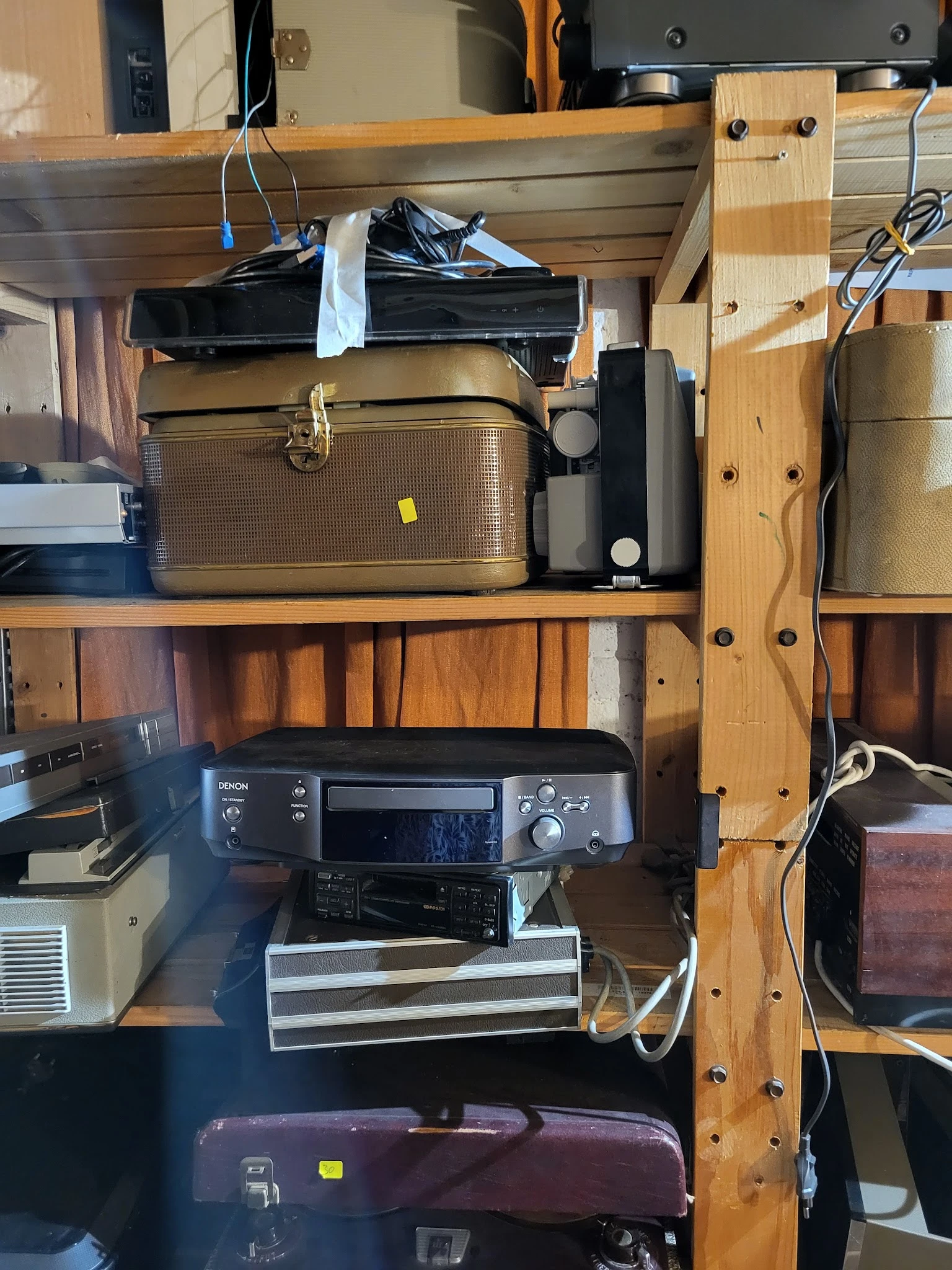 Tweedehands elektronische apparatuur op houten stellingen, waaronder een goudkleurige koffer, een printer en andere elektroni