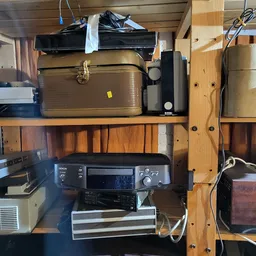 Tweedehands elektronische apparatuur op houten stellingen, waaronder een goudkleurige koffer, een printer en andere elektroni