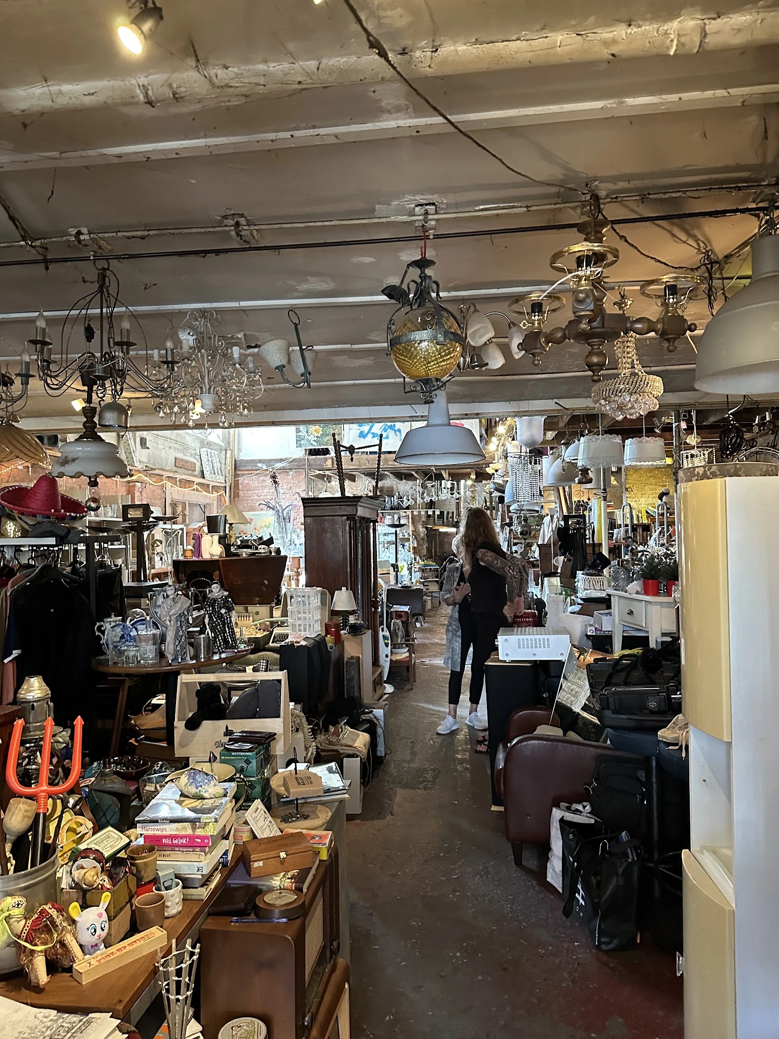 Binnenaanzicht van kringloopwinkel met hanglampen, meubels, kleding en diverse spullen op stellingen en tafels